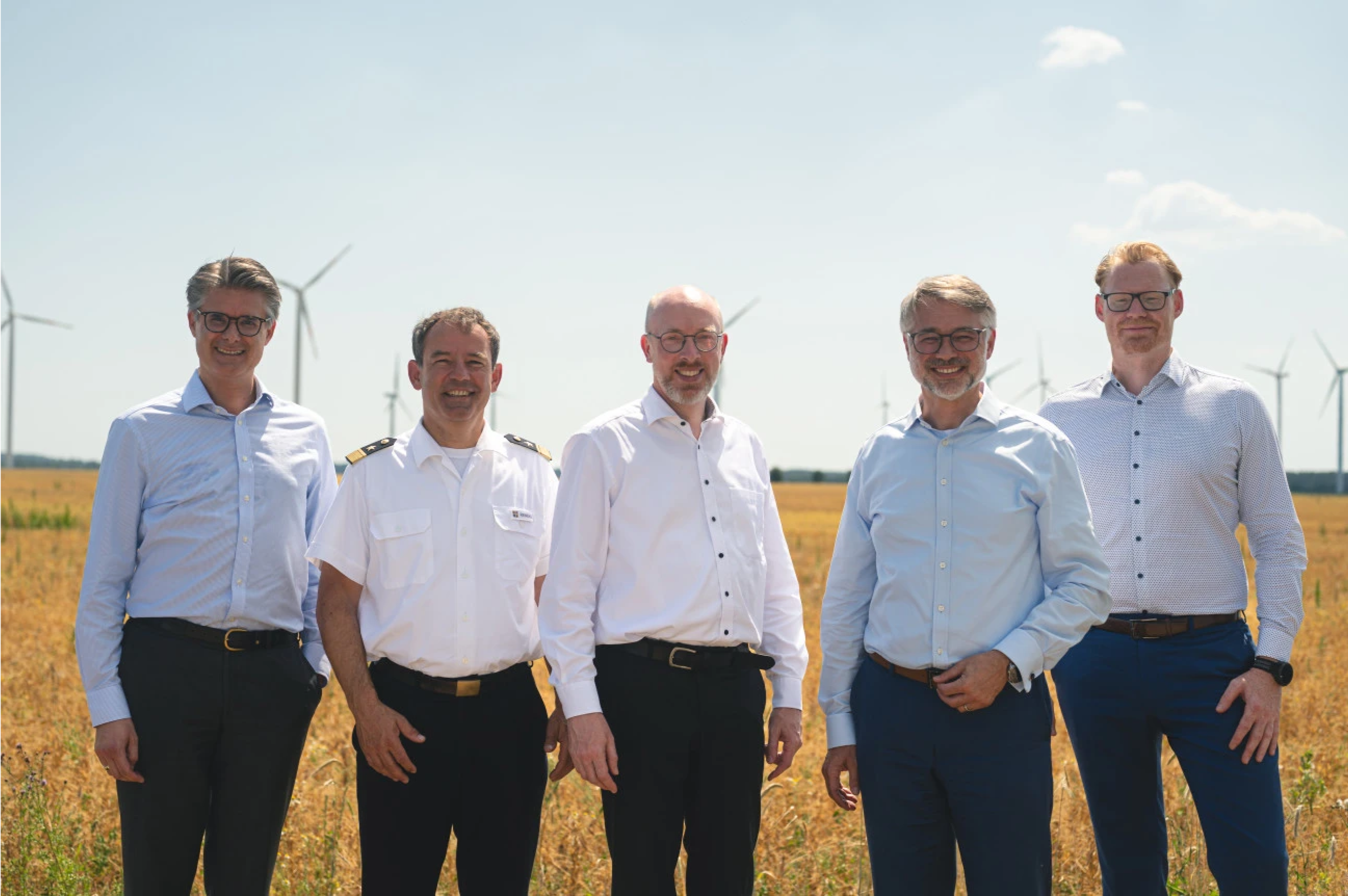 Fünf Männer stehen lächelnd in einem Feld mit Windkraftanlagen im Hintergrund.