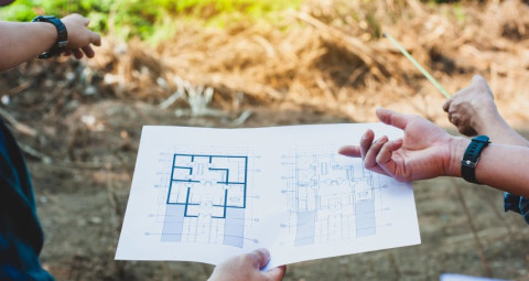 Personen halten einen Bauplan und zeigen auf eine Baustelle im Hintergrund.
