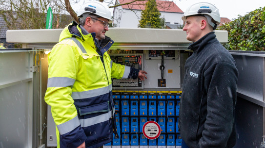 Zwei Fachkräfte in Schutzkleidung arbeiten gemeinsam an einem elektrischen Schaltkasten im Freien.