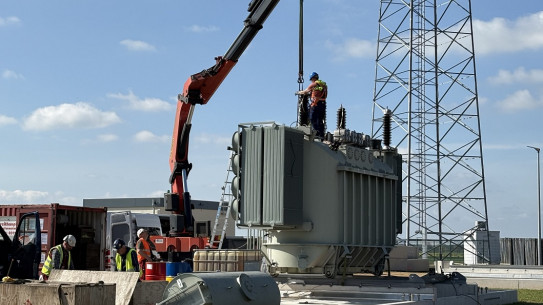 Arbeiter heben einen Transformator mit einem Kran auf eine Plattform. Im Hintergrund ist ein Strommast zu sehen.