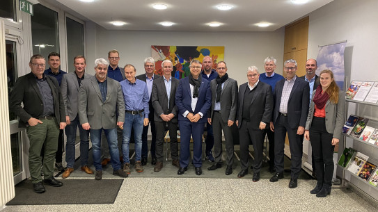 Eine Gruppe von 17 Personen steht in einer Lobby, im Hintergrund Farbenfrohes Kunstwerk und Informationsstände.