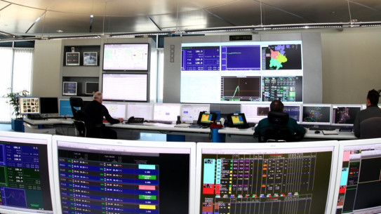 Blick in ein Kontrollzentrum mit mehreren Monitoren und zwei Personen, die Daten auswerten.