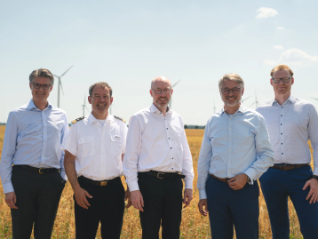 Fünf Männer stehen lächelnd in einem Feld mit Windkraftanlagen im Hintergrund.