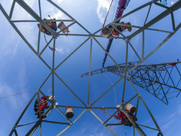 Blick von unten auf einen Strommast mit Arbeitern und einem Kran im Einsatz unter blauem Himmel.