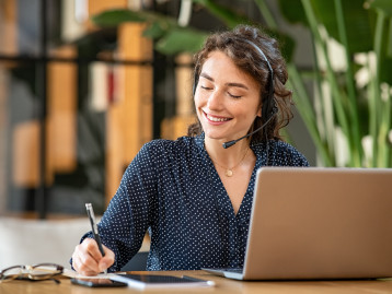 Frau mit Headset lächelt beim Arbeiten am Laptop, notiert etwas auf einem Block. Pflanzen im Hintergrund.