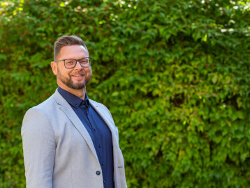 Mann mit Brille und grauem Blazer lächelt vor einer grünen Pflanzenwand.