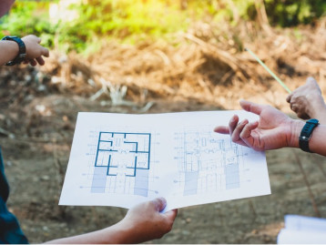 Personen halten einen Bauplan und zeigen auf eine Baustelle im Hintergrund.