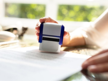 Hand hält einen Stempel über einem Dokument auf einem Tisch mit Fensterblick im Hintergrund.
