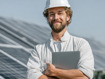 Mann mit Helm und Laptop steht vor Solarpanelen, lächelt und trägt eine helle Jacke.