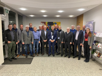 Eine Gruppe von 17 Personen steht in einer Lobby, im Hintergrund Farbenfrohes Kunstwerk und Informationsstände.