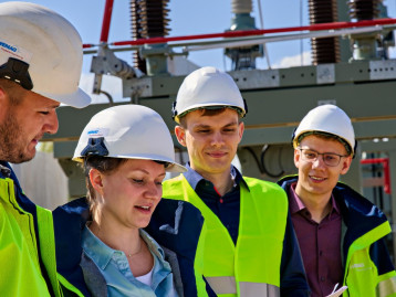 Gruppe von vier Personen in Schutzhelmen und Warnwesten bespricht technische Details vor einer Industrieanlage.