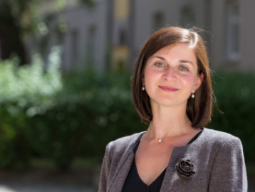 Porträt einer Frau mit braunen Haaren, die in einem hellen Umfeld steht und einen grauen Blazer trägt.