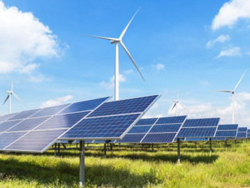 Windkraftanlagen hinter Solarpanels auf einer Wiese bei blauem Himmel und weißen Wolken.