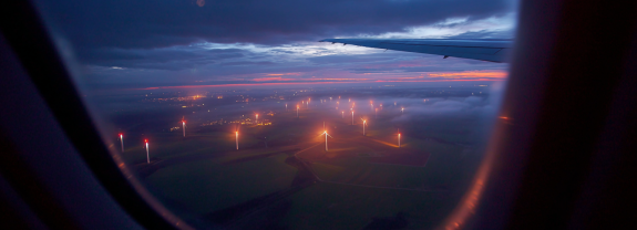 Blick aus einem Flugzeugfenster auf Windkraftanlagen und eine städtische Landschaft bei Sonnenuntergang.