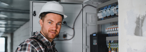 Ein Elektriker mit weißem Schutzhelm und Arbeitskleidung steht vor einem geöffneten Schaltschrank und arbeitet an elektrischen Komponenten; Kabel und Sicherungen sind sichtbar