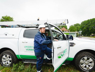 Mann in Arbeitskleidung steht neben einem weißen Transportfahrzeug mit Leitern auf dem Dach inmitten einer grünen Umgebung.