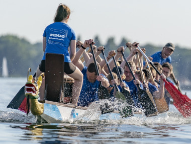Wettkampf im Drachenboot mit mehreren Paddlern auf einem Gewässer, im Hintergrund Segelboote.