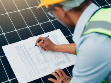 Person in gelber Schutzbekleidung schreibt auf ein Dokument, während sie einen Solarpanel-Hintergrund betrachtet.