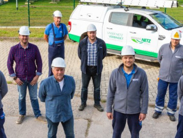 Eine Gruppe von acht Personen in Arbeitskleidung und Helmen steht auf einem Hof, im Hintergrund ein Nutzfahrzeug.