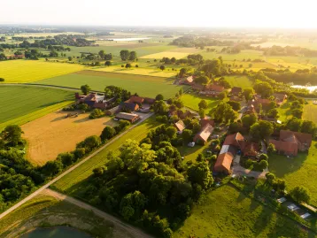 Luftaufnahme eines Dorfes umgeben von Feldern, Wäldern und einer Wasserfläche bei Sonnenuntergang.
