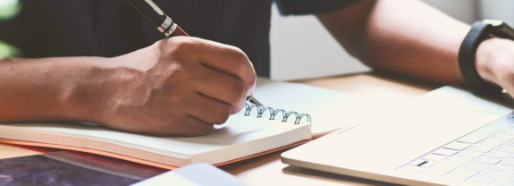 Eine Hand schreibt mit einem Stift in ein geöffnetes Notizbuch auf einem Tisch.