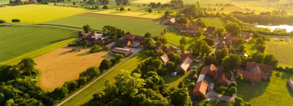 Luftaufnahme eines Dorfes umgeben von Feldern, Wäldern und einer Wasserfläche bei Sonnenuntergang.