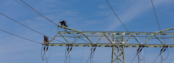 Arbeiter führen Wartungsarbeiten an einem Hochspannungsmast unter blauem Himmel durch.
